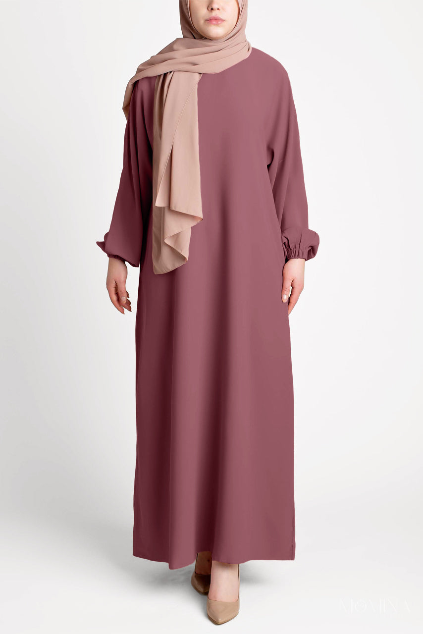 Woman wearing a long mauve dress with a beige hijab on a white background